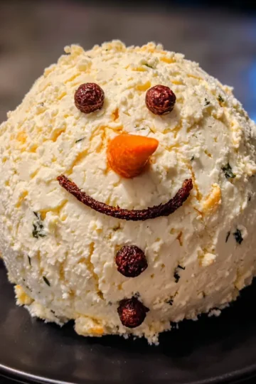 A festive Christmas snowman cheese ball recipe, beautifully crafted with a carrot nose and herb specks, presented on a dark plate.