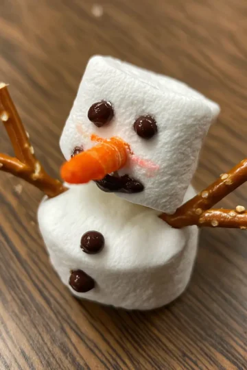 A charming edible christmas snowman marshmallow recipe creation, featuring stacked marshmallows, pretzel arms, candy eyes, a carrot nose, and chocolate buttons, resting on a wooden surface.