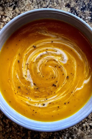 Overhead view of a creamy butternut squash soup recipe garnished with a cream swirl and black pepper on a speckled countertop.
